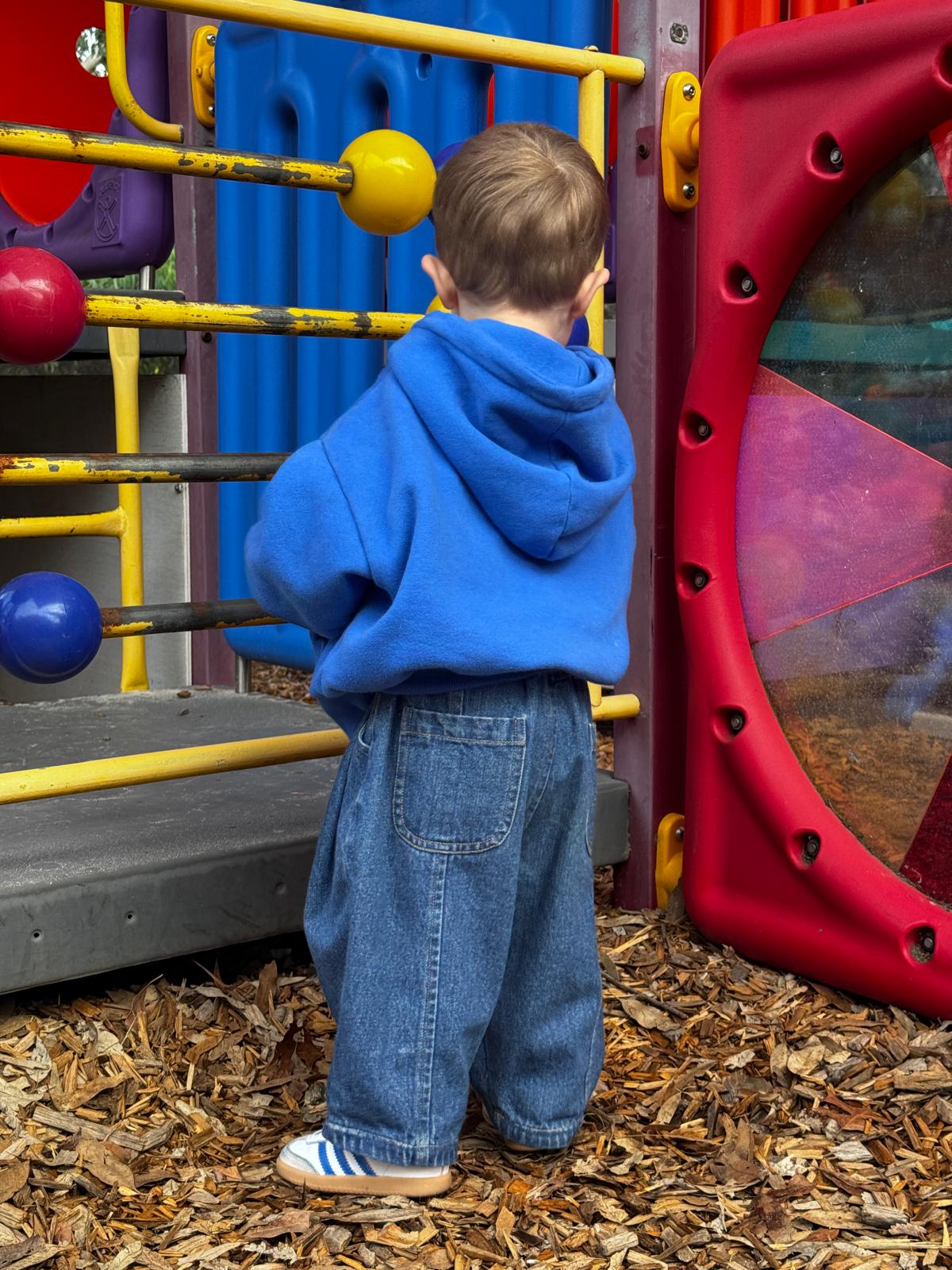 Child in a blue hoodie and ruhi jeans standing on a playground with colorful equipment.