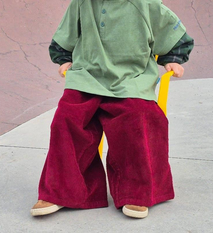 Child wearing a green plaid ollie double-sleeve shirt and red corduroy blaze pants standing on a concrete surface with a pink wall in the background.