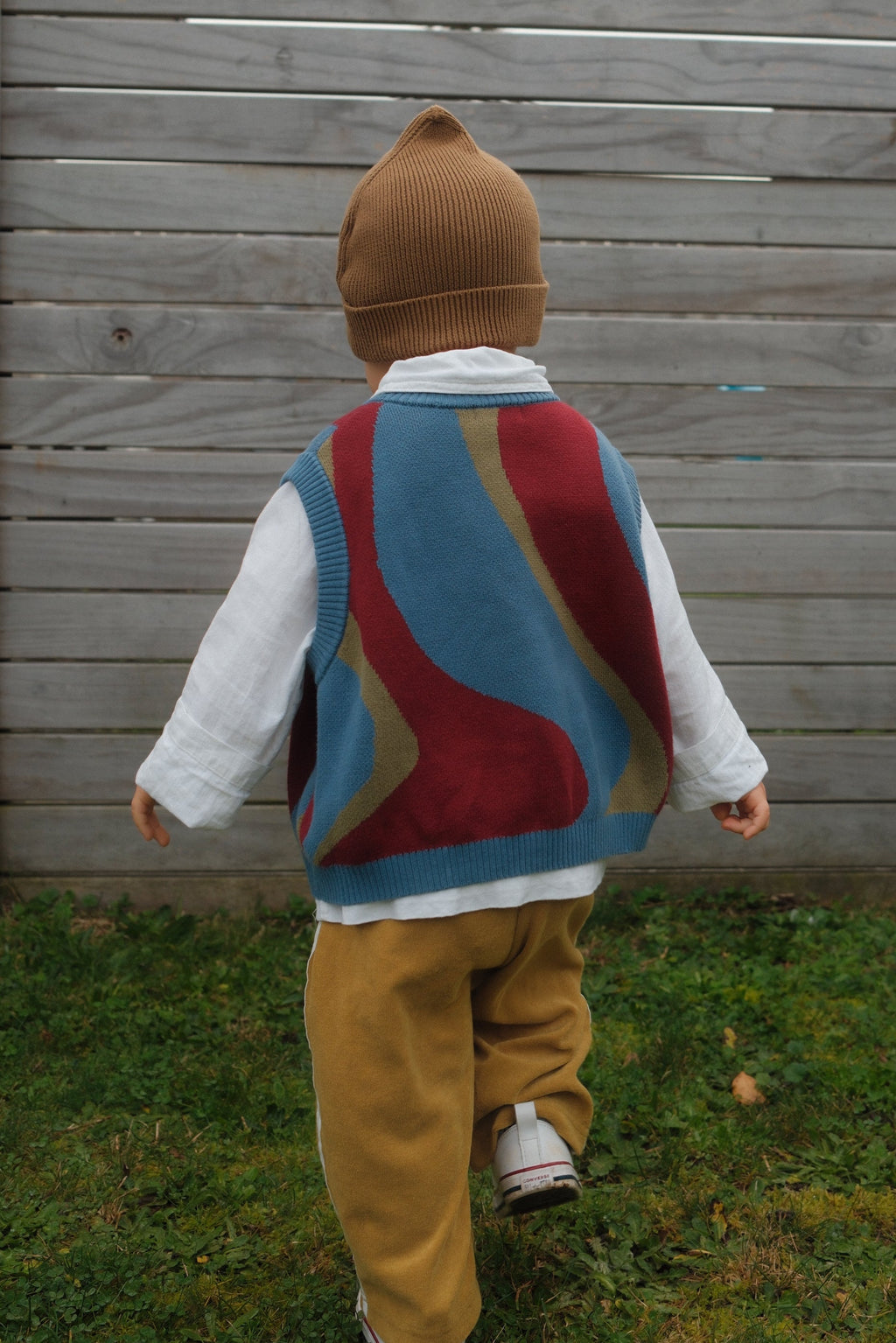 A child viewed from behind wearing a brown knit beanie, a colorful johnny sweater vest in cherry ice swirl over a white long-sleeve shirt, mustard yellow pants, and white sneakers, standing near a gray wooden fence on green grass.
