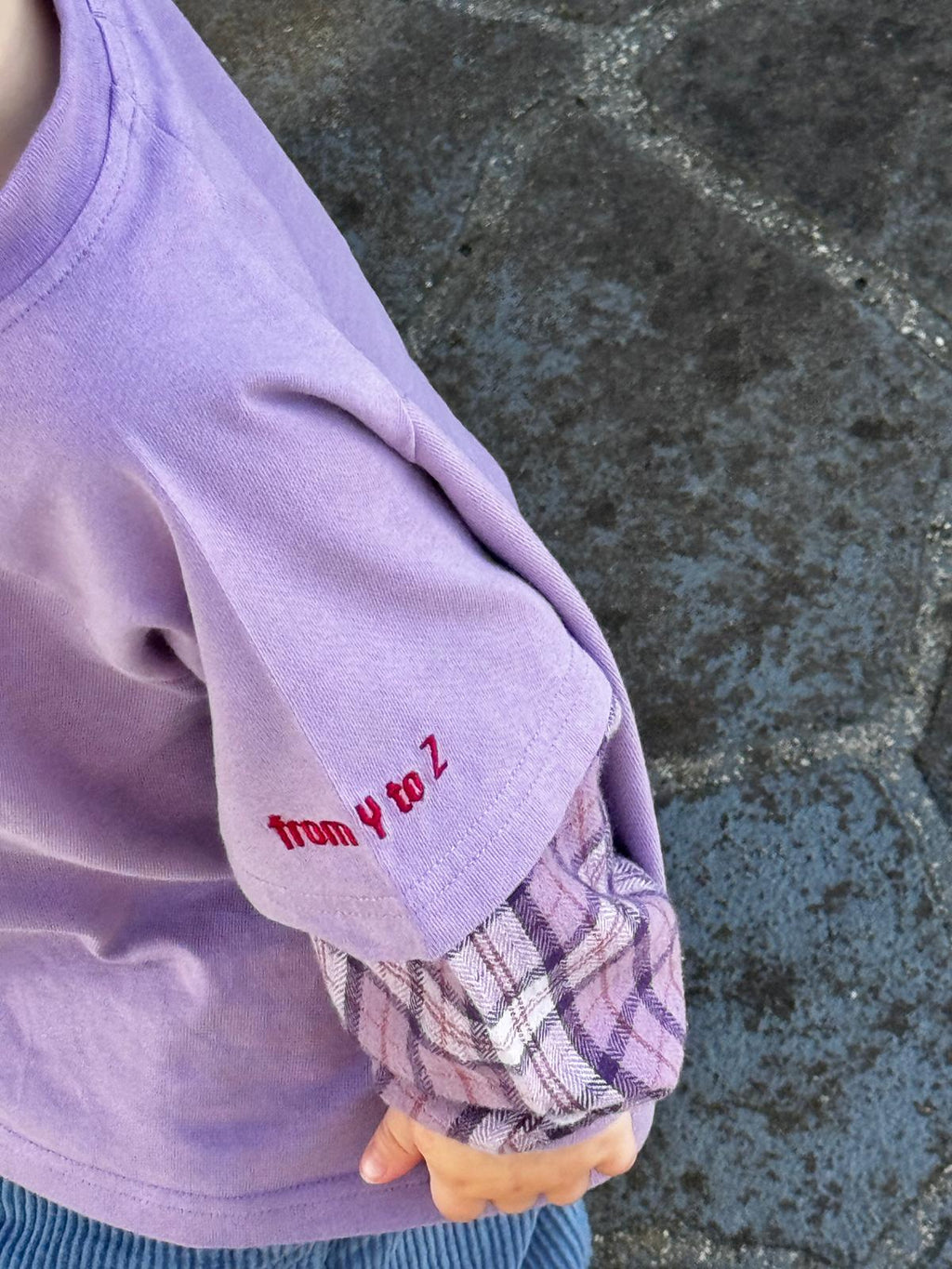 Close-up of a child's arm wearing a purple sleeve with plaid cuffs against a stone pavement background.