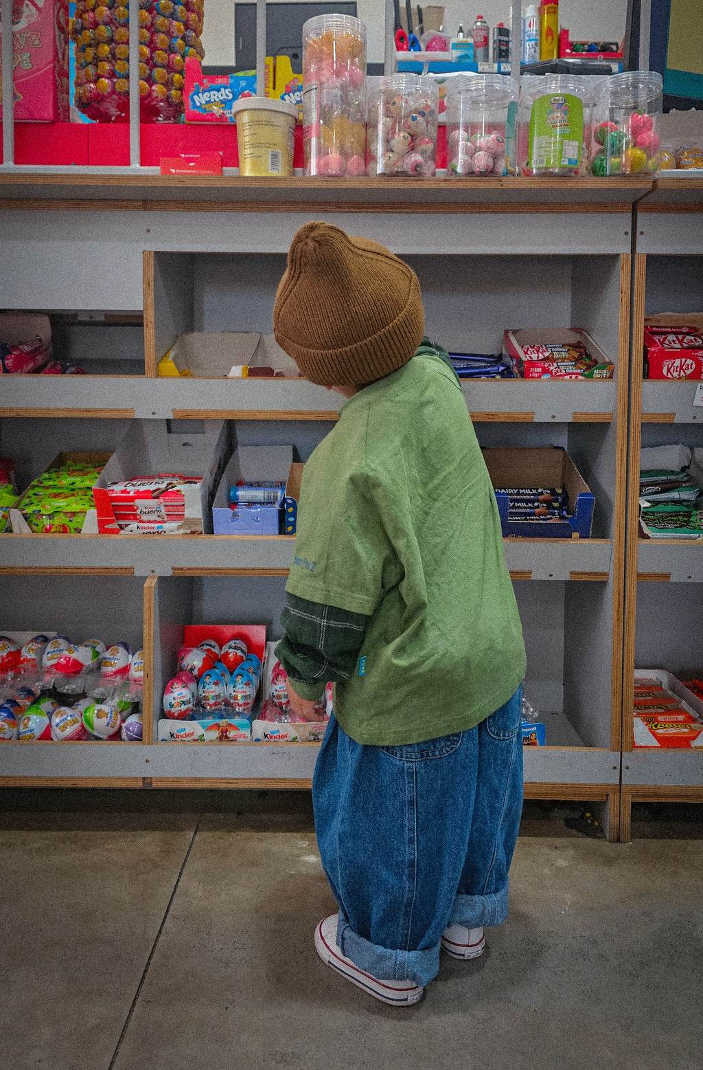 Child in a green hunter plaid tee and brown beanie standing in front of a store shelf with snacks.