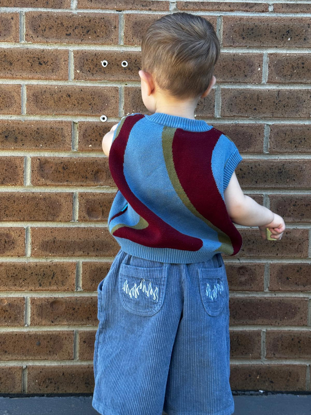 Child wearing a colorful vest and blue corduroy wide leg pants against a brick wall