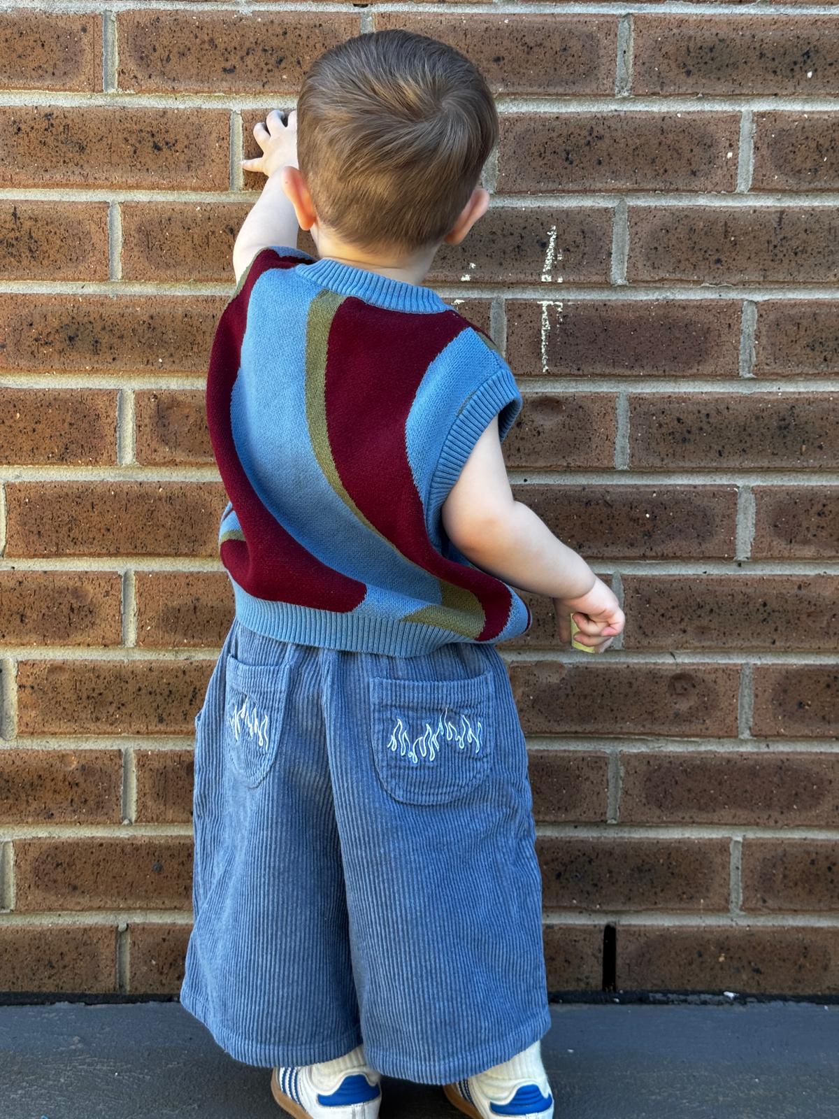 Child wearing a colorful johnny sweater vest and blue pants against a brick wall.