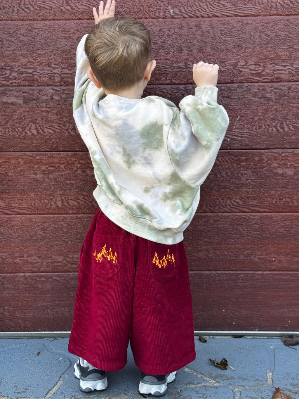 Child wearing a tie-dye hoodie and red corduroy pants with flame designs against a wooden wall.