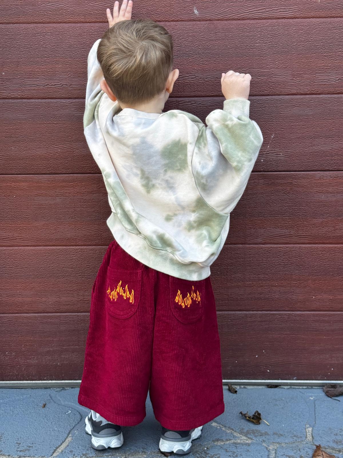 Child wearing a tie-dye hoodie and red corduroy pants with flame designs against a wooden wall.